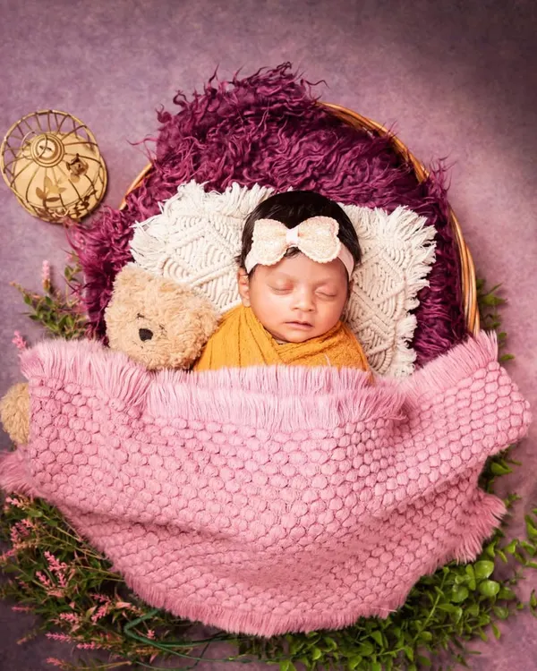 A perfect little bundle sleeping soundly in a basket. The baby is wrapped in a warm mustard-colored cloth and tucked in with a pink blanket, accompanied by a tiny teddy bear.