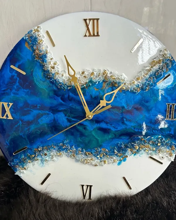 A close-up of a geode-inspired wall clock. The deep blue resin flows beautifully into the white, with shimmering gold crystals creating a natural, luxurious divide. The gold Roman numerals complete this elegant timepiece.