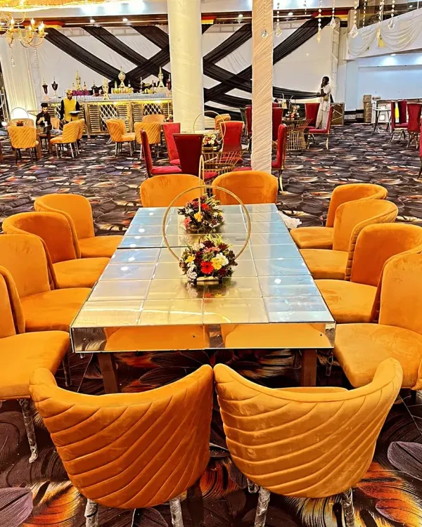 This is a long dining table setup for guests, featuring a mirrored tabletop that reflects the decor. The comfortable orange velvet chairs add a pop of color to the event space.