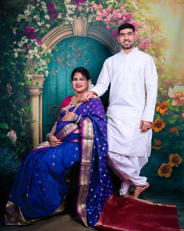 A beautiful couple portrait from a traditional maternity shoot in our studio. The mother-to-be is seated gracefully in a royal blue saree, complemented by a floral archway backdrop.