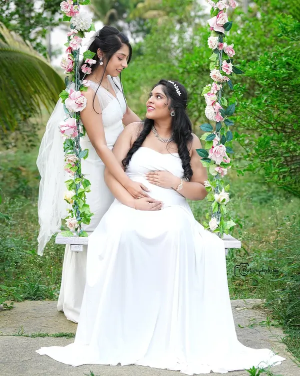 A beautiful moment between sisters or friends celebrating the upcoming arrival. Both are dressed in elegant white gowns, sharing a look of love and support.