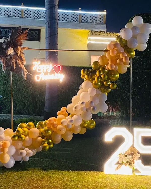 An elegant outdoor setup for a 25th anniversary party. This features a diagonal balloon garland, a custom neon sign, dried floral arrangements, and a large illuminated '25'.