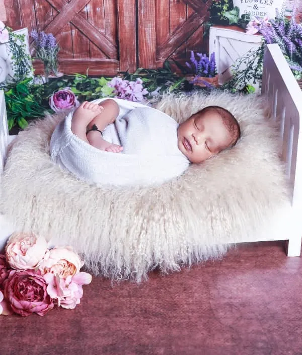 A classic newborn pose with the baby swaddled and sleeping peacefully in a miniature white bed. The rustic floral backdrop adds a touch of warmth and charm.