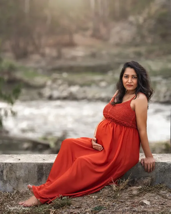 An expecting mother in a flowing red dress sits peacefully by a flowing stream. This outdoor portrait combines the raw beauty of nature with a serene, contemplative mood.