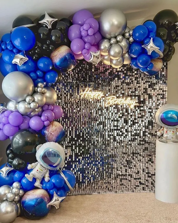 An out-of-this-world space theme birthday party. This setup features a silver shimmer wall, a planet-themed balloon garland in blue, purple, and silver, and astronaut props.