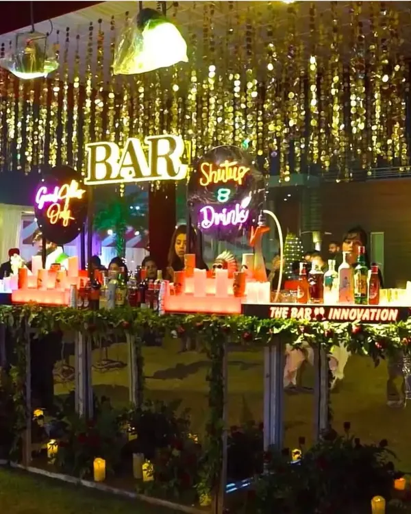 A stunning bar setup for a cocktail night. The illuminated bar is decorated with neon signs, greenery, and candles, with a ceiling of hanging gold tassels creating a glamorous look.