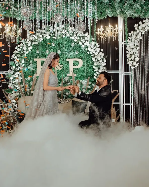 A fairytale proposal moment during the engagement, with the groom on one knee, surrounded by theatrical smoke and a beautiful floral backdrop.
