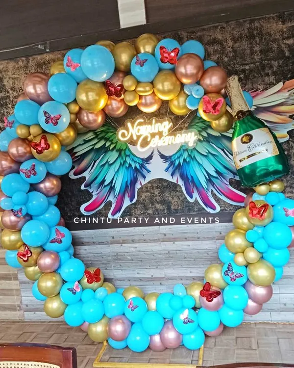 A vibrant naming ceremony balloon ring in blue, gold, and rose gold. The design features a neon sign with angel wings and butterfly accents.