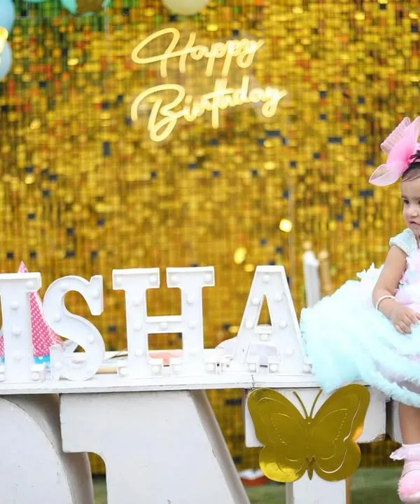 A complete setup for a first birthday photoshoot, featuring a sparkly gold backdrop, a light-up name sign, and pastel balloons. The birthday girl is dressed in a beautiful party dress to complete the look.