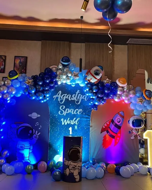 This is the hero item. A spectacular space-themed birthday bash for a first birthday. This setup includes a custom "Agastya Space World" backdrop, a full organic balloon arch in blues and silver, astronaut cutouts, and a large light-up number '1' to mark the big milestone.