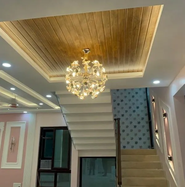 A luxurious villa interior featuring a stunning crystal chandelier hanging from a wooden false ceiling panel. The multi-level ceiling design and wall molding add depth and grandeur to the space.