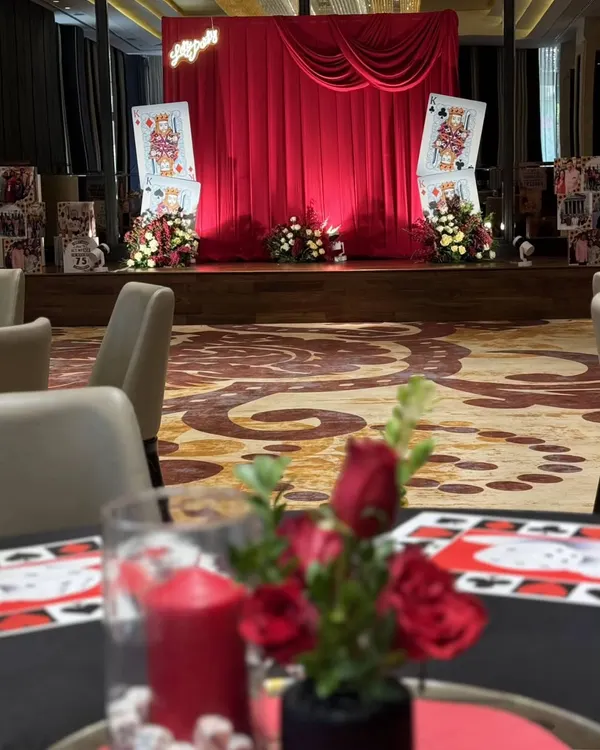 A view from a guest's table at our casino-themed event. The red rose centerpiece in the foreground complements the dramatic red draping of the main stage in the background, creating a cohesive and luxurious atmosphere throughout the room.
