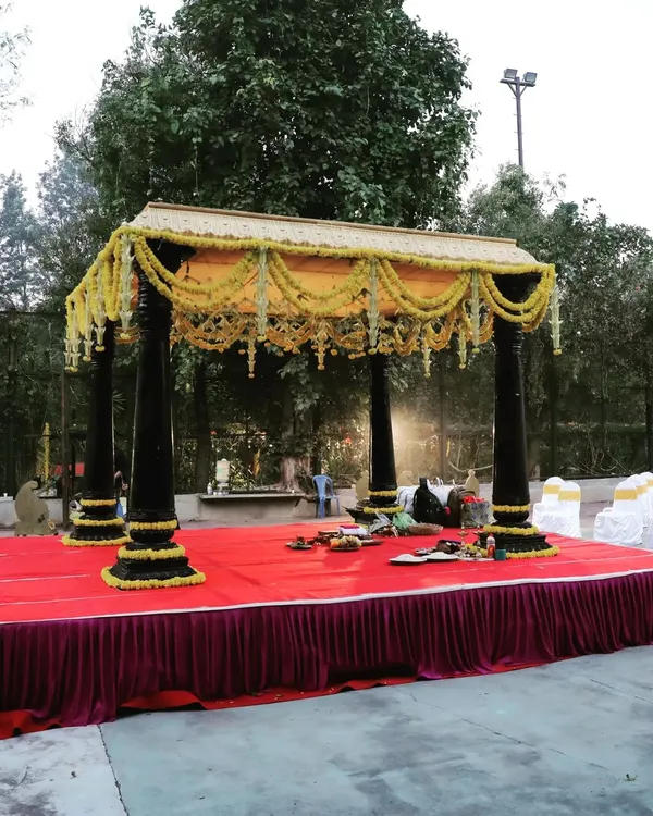 This outdoor wedding mandap combines sturdy, dark pillars with delicate garlands of yellow flowers. The setup is placed on a raised red platform, creating a defined and sacred space for the wedding ceremony.