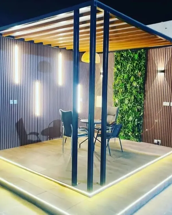 This is how we transform an empty terrace into a sophisticated evening retreat. The design combines a sleek black pergola, warm wood-look paneling, and vertical tube lighting with a lush artificial green wall, all illuminated by elegant floor-level LEDs.