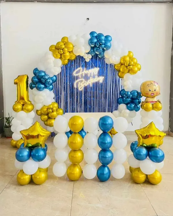 A classic first birthday setup with a blue, yellow, and white balloon arch. This decoration includes a "Happy Birthday" neon sign against a blue fringe curtain and a large number '1' foil balloon.