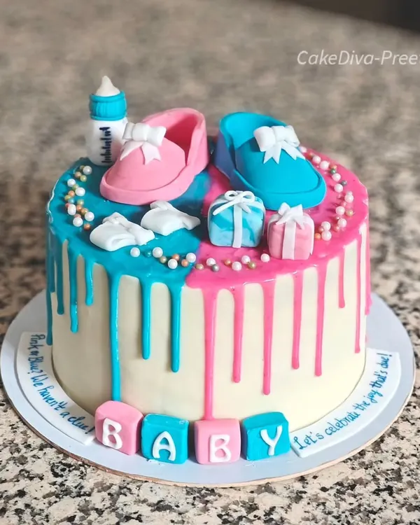 Pink or blue, the surprise is true! This gender reveal cake features pink and blue chocolate drips and is topped with cute, handmade fondant baby shoes, a bottle, and gift boxes.