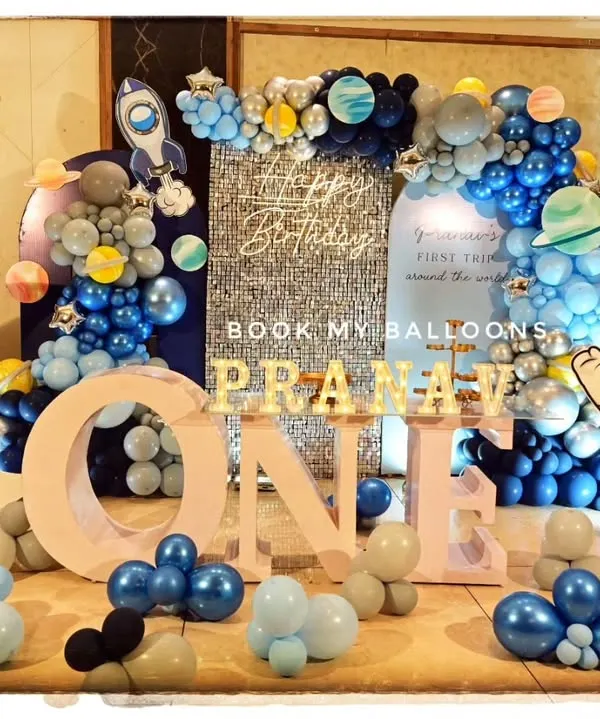 A spectacular space-themed first birthday party, featuring a large moon prop, rocket cutouts, an astronaut figure, and a stunning balloon galaxy arch.