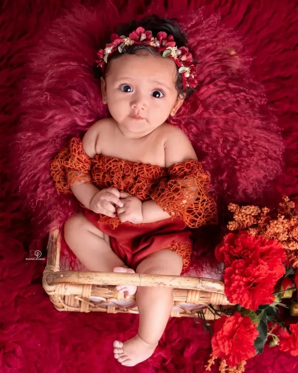 A striking portrait of a three-month-old baby girl. Dressed in a lacy red outfit with a matching floral headband, she sits in a small basket against a rich, textured red background.