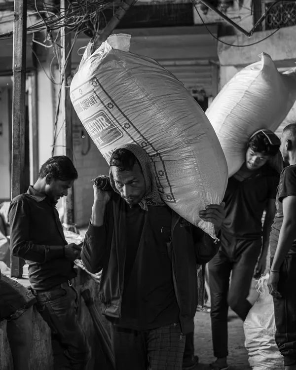 A laborer carries a heavy sack through the crowded lanes of Khari Baoli spice market. This black and white documentary shot highlights the grit and hard work that defines daily life here.