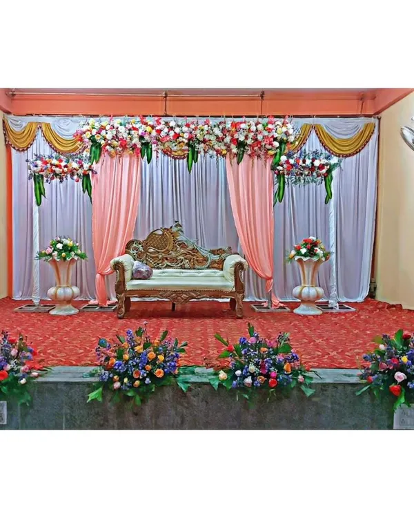 This is our classic stage decoration for a formal event like a reception or engagement. The setup includes a beautiful floral valance, elegant pink and white drapes, a regal lounge sofa, and matching flower arrangements on urns.