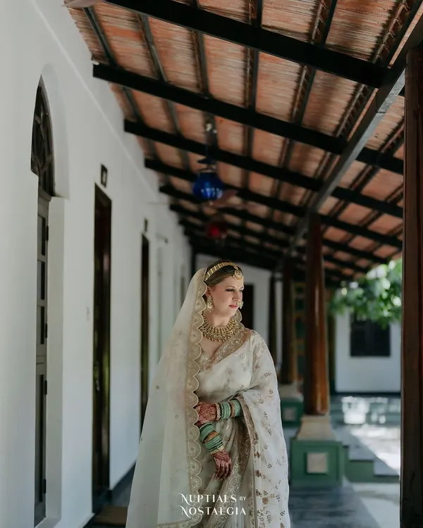 A bride in a stunning white saree stands in a sunlit corridor, the repeating pillars creating a beautiful, elegant frame for her portrait.