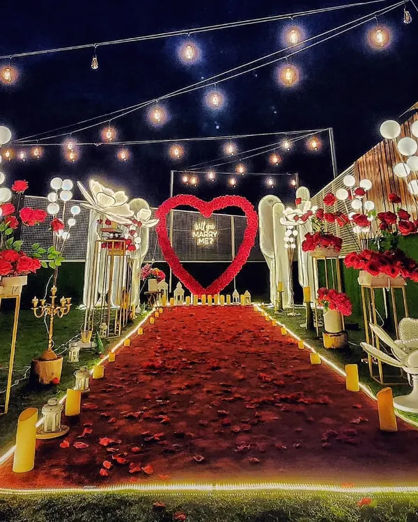 A nighttime view of a spectacular proposal setup. The combination of a red rose petal aisle, a glowing heart arch, and dozens of LED candles creates an intensely romantic and picture-perfect scene against the dark sky.