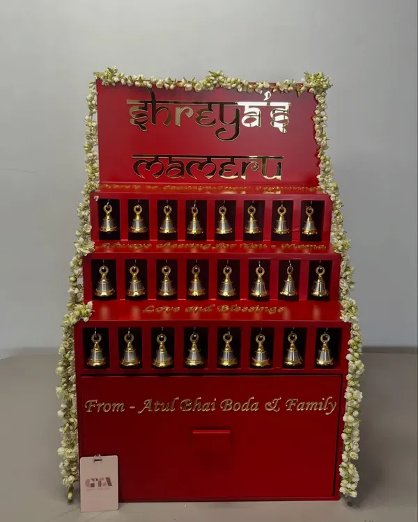 This custom-built structure was designed for a 'Mameru' ceremony, a cherished tradition of gifting from the maternal family to the bride. The vibrant red is accented with gold lettering in Hindi and delicate jasmine garlands, with each niche holding a small, symbolic bell.