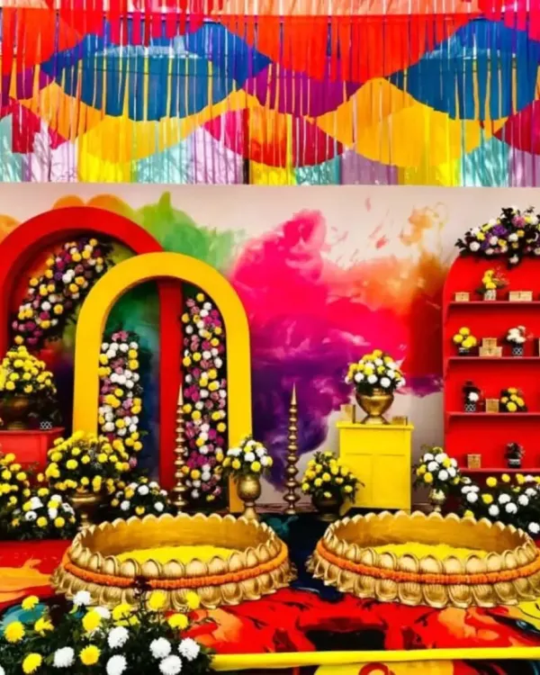 A complete Haldi ceremony stage featuring a backdrop of colorful powder explosions, vibrant floral arrangements in yellow and purple, and traditional brass urlis for the rituals. The multi colored ribbon ceiling adds to the festive feel.
