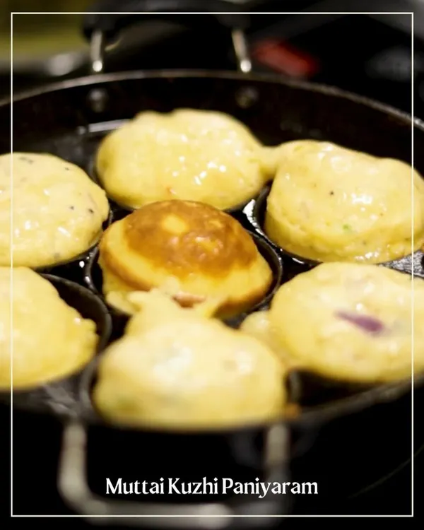 A close-up of Muttai Kuzhi Paniyaram sizzling in a traditional cast-iron pan. These savory Chettinad-style egg and rice batter puffs are cooked until golden brown and fluffy.