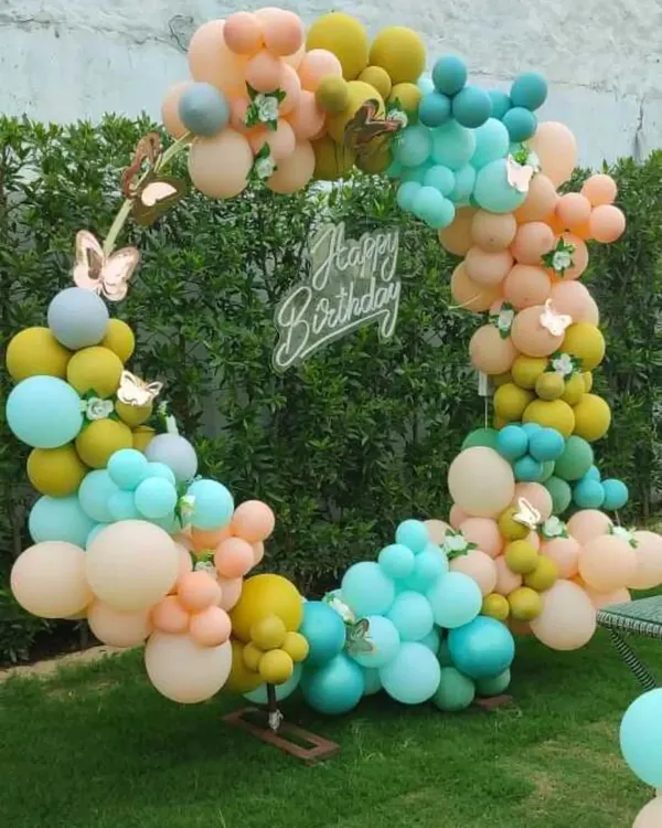 A beautiful pastel balloon ring set up in a garden. This outdoor decoration, adorned with delicate butterflies, creates a whimsical and enchanting photo backdrop, perfect for a daytime birthday party.