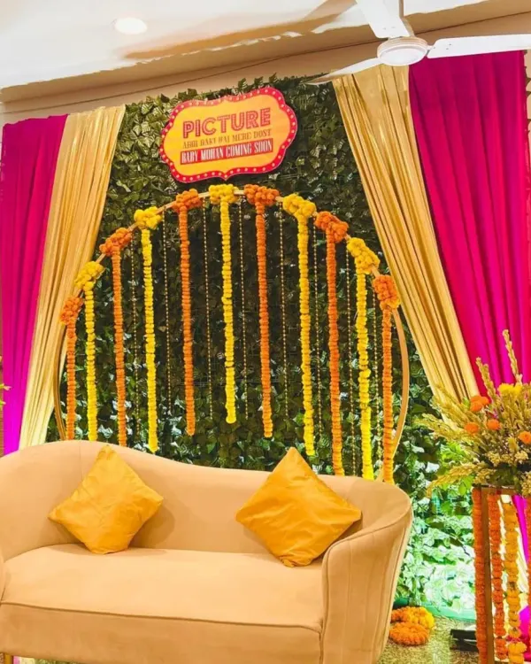 A vibrant and filmy Godh Bharai setup. We used a lush green wall as a base, adorned with an arch of fresh marigolds and bright pink and gold drapes for a festive, celebratory feel.