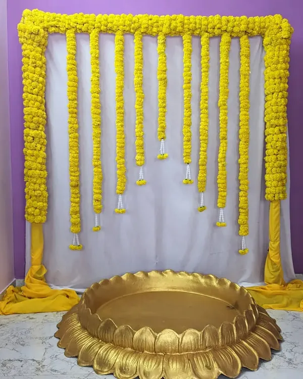 A classic Haldi ceremony backdrop. This simple and elegant design features a frame draped in yellow fabric with hanging marigold garlands, paired with a large golden lotus-shaped bowl (urli).