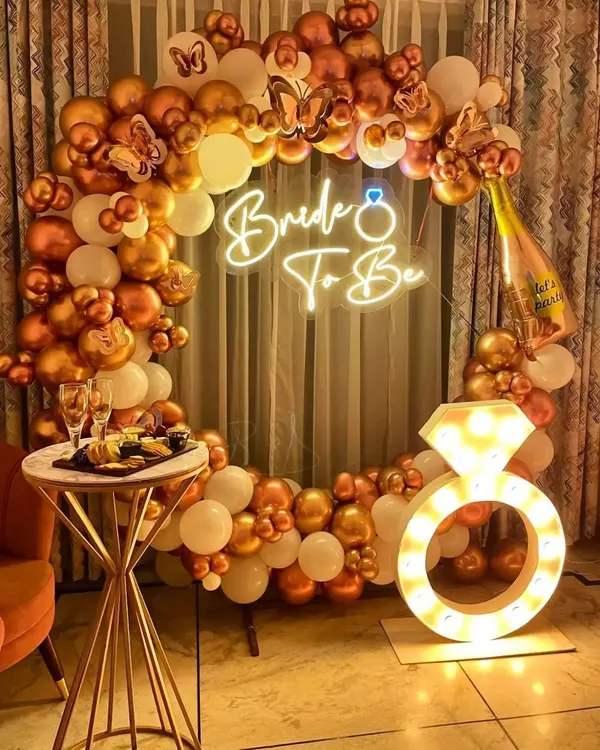 A glamorous "Bride To Be" setup perfect for a bachelorette party. This decoration features a circular balloon arch in white and chrome gold, a neon sign, butterfly accents, and a large light-up diamond ring prop.
