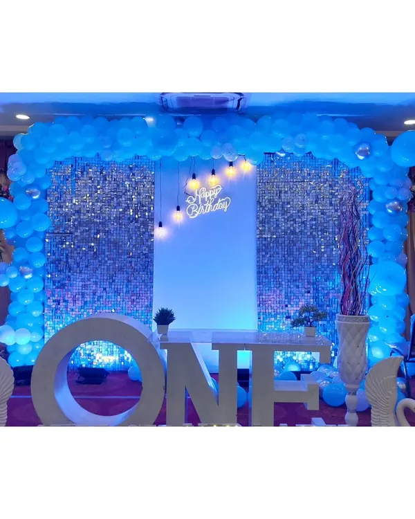 A stunning setup for a first birthday, featuring a large light-up 'ONE' sign as the centerpiece. The backdrop is a silver shimmer wall framed by a blue balloon arch, creating a cool, celebratory glow for the big milestone.