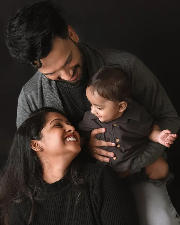 A classic family portrait that radiates warmth and connection. The dark, coordinated outfits create a timeless, fine-art feel, drawing all the attention to the loving expressions and smiles shared between the parents and their baby.