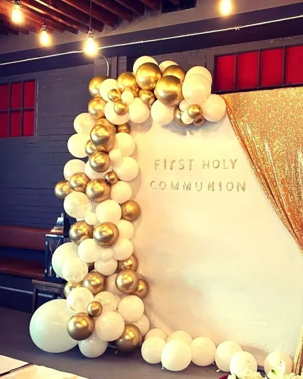 An elegant First Holy Communion decoration featuring a white and gold balloon arch against a white backdrop with a gold sequin curtain. This creates a sophisticated and celebratory setting for the milestone.