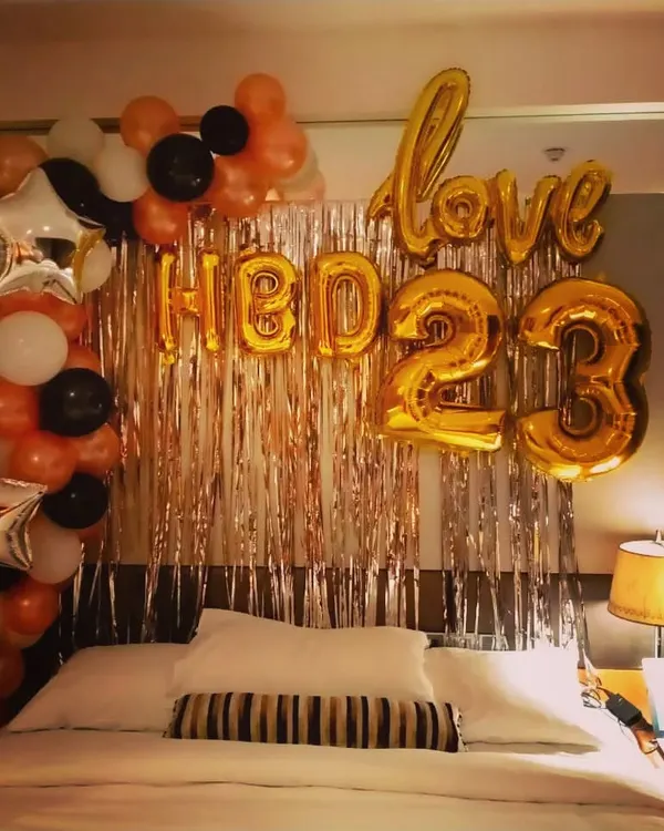 A classic bedroom birthday surprise for a partner, celebrating their 23rd birthday. This decoration features "HBD 23" and "love" foil balloons against a rose gold shimmer curtain, framed by a balloon arch in black, orange, and white.