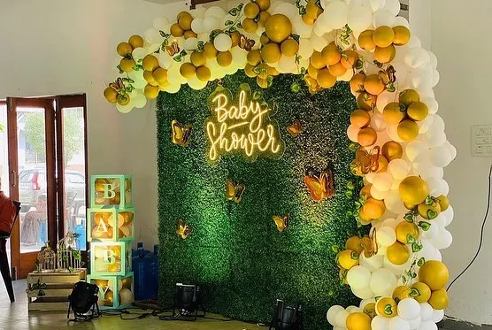 A stylish baby shower decoration featuring a lush green foliage wall with a 'Baby Shower' neon sign. The wall is framed by a white and yellow balloon arch with butterfly details.