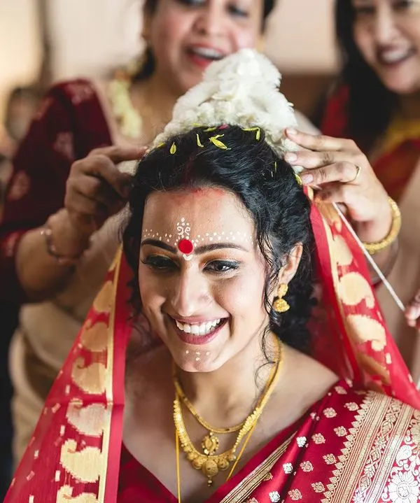 The joy of a Bengali bride. This look features the traditional chandan design on the forehead, a bold red bindi, and classic gold jewelry, capturing the essence of a Bengali wedding.