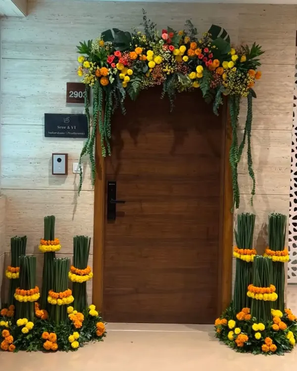 This door decoration offers a modern take on the traditional marigold. A lush arch is accented with hanging greenery and creative floral bundles at the base, creating a stylish and welcoming entrance.