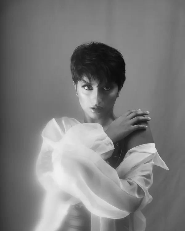 A soft-focus, ethereal black and white portrait of a woman with a pixie cut, wrapped in a sheer white blouse. The dreamy glow gives this studio headshot an artistic and gentle quality.