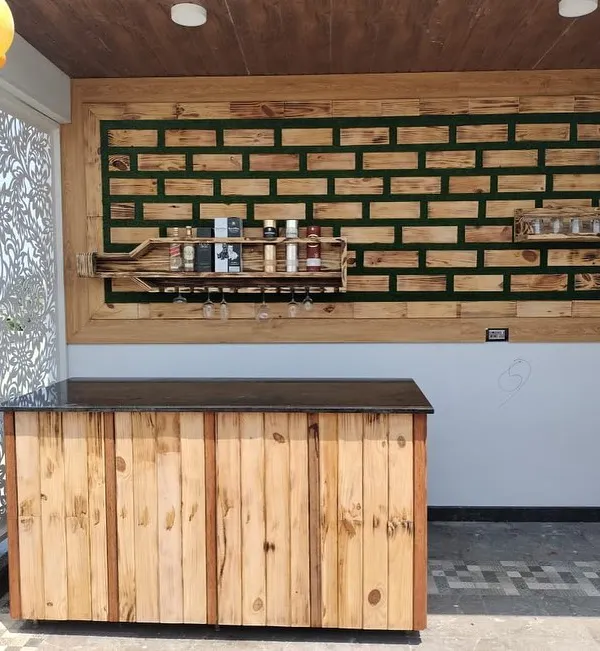 A complete view of a custom bar installation on a covered terrace. The setup includes a rustic wood-paneled counter with a dark top, paired with the signature bottle-shaped wall shelf and decorative paneling.