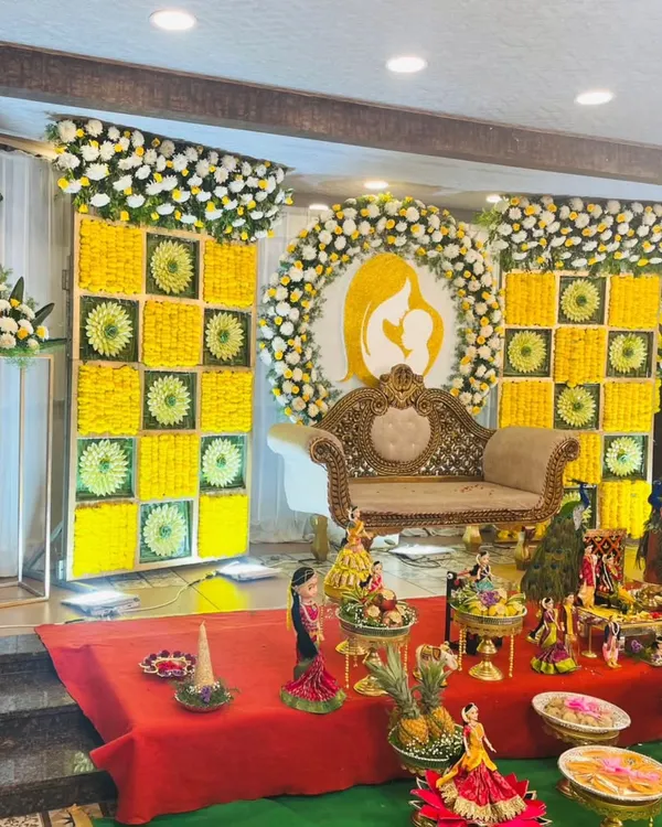 A stunning stage for a traditional baby shower, or Seemantha. The backdrop features intricate panels of yellow and white flowers with a central silhouette of a mother and child, creating a meaningful and beautiful setting.