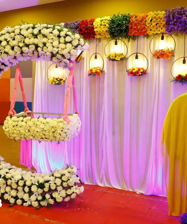 A stunning cradle ceremony setup featuring a crescent-shaped arch made of white flowers. The hanging cradle, also adorned with flowers, creates a beautiful focal point, while colorful lanterns add a festive touch.