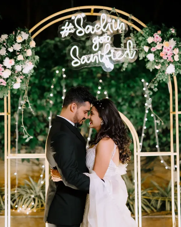 A romantic moment for the newly engaged couple, framed by a modern golden arch decorated with pastel flowers and a custom neon sign. The fairy lights and lush green background add a touch of magic.