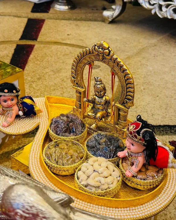 A charming dry fruit platter for a baby naming ceremony. The centerpiece is a golden swing with a baby Krishna idol, surrounded by bowls of cashews and raisins and cute baby dolls.