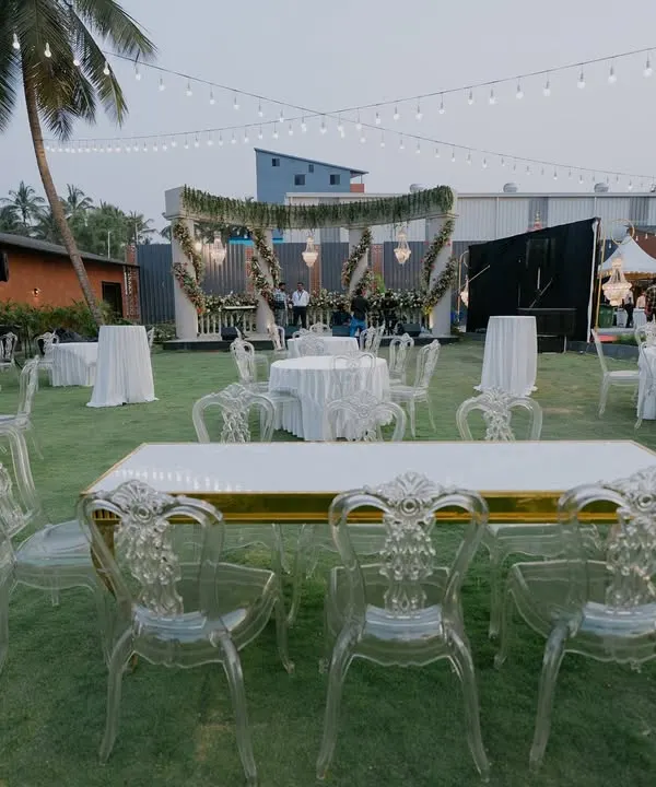 An elegant outdoor reception setup on a lush green lawn. The combination of modern ghost chairs, classic chandeliers, and string lights creates a sophisticated yet relaxed atmosphere.