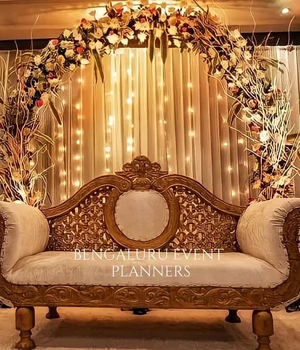I designed this elegant stage for a wedding reception, featuring a classic carved sofa set against a backdrop of warm fairy lights. The setup is framed by a lush floral arch, creating a beautiful and timeless focal point for the couple.