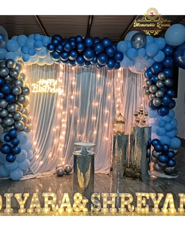 A beautiful birthday setup with a blue and silver balloon arch over a backdrop of sheer curtains and fairy lights. The names are highlighted with marquee letters.