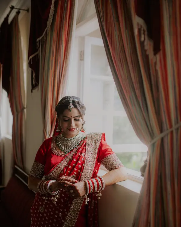 An elegant bridal portrait of the bride in her red saree, captured in beautiful natural light by a window. These quiet, reflective moments before the ceremony make for timeless photos.